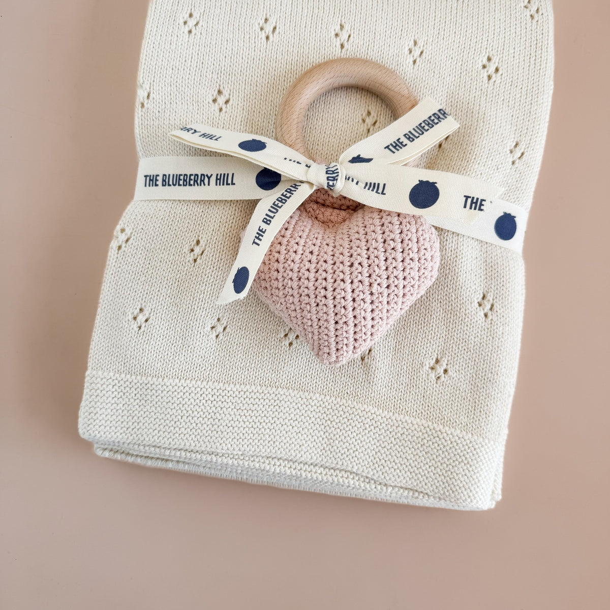 Pink heart-shaped teething ring with a wooden handle on a folded blanket, branded 'The Blueberry Hill'.