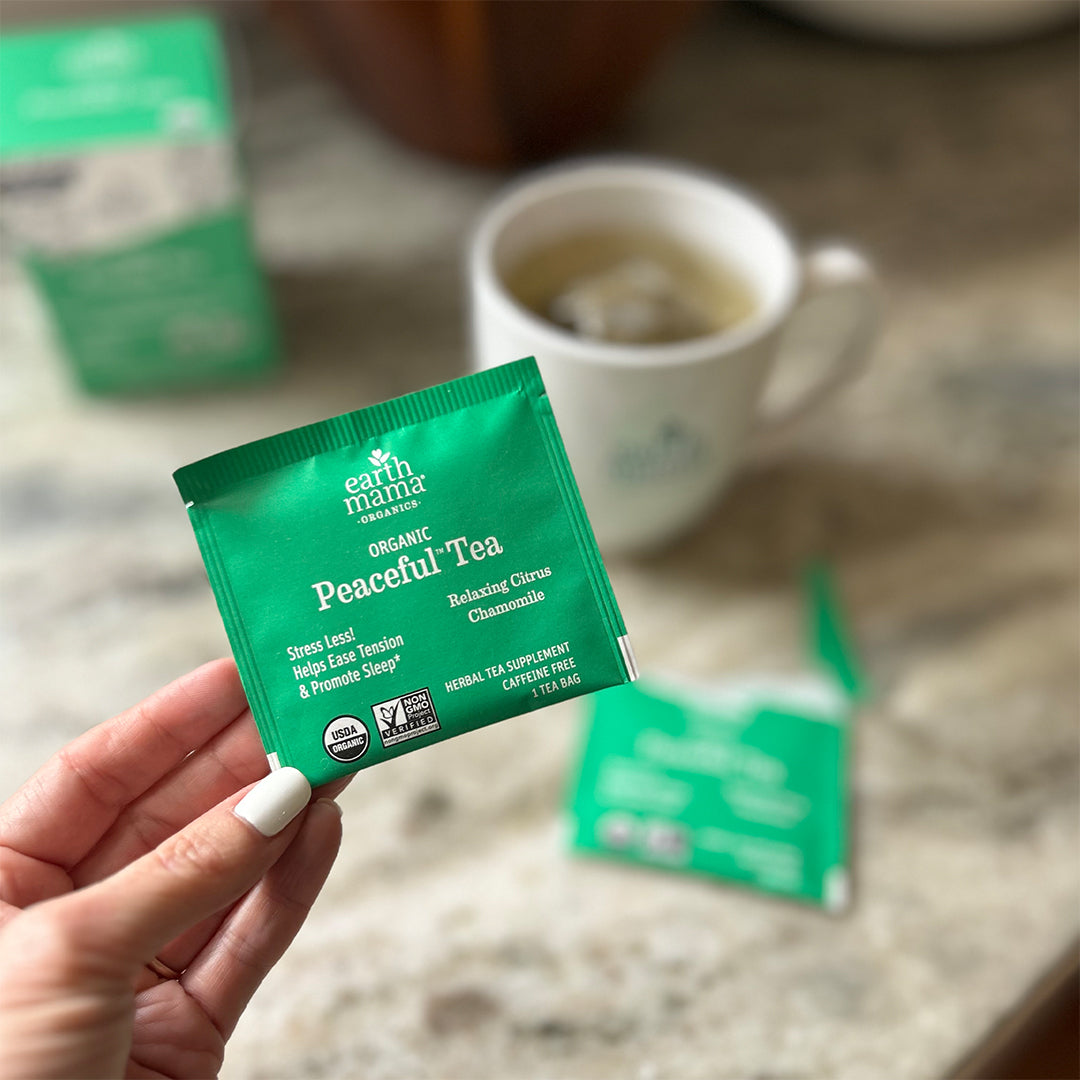 Hand holding an Earth Mama Organic Peaceful Tea packet with a blurred cup of tea in the background.