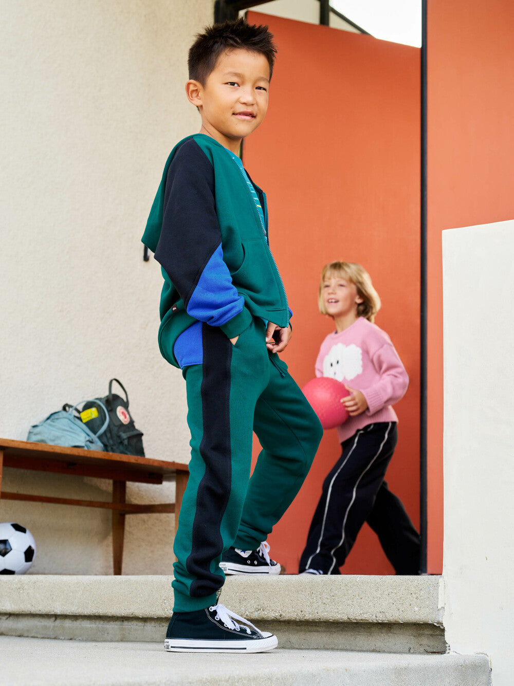 Child in green and navy tracksuit standing on steps with another child in pink shirt in the background.