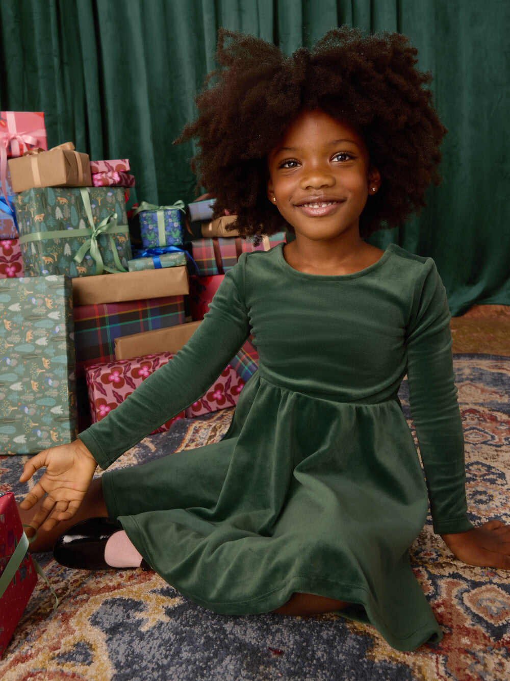 Child in a green dress sitting on the floor with Christmas presents in the background