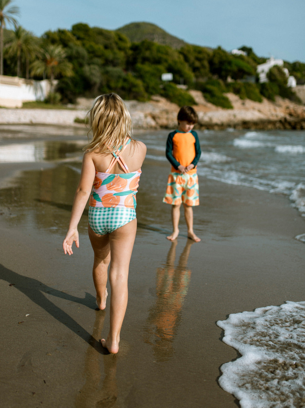 Two children on a beach with one walking towards the water and the other holding an orange float.