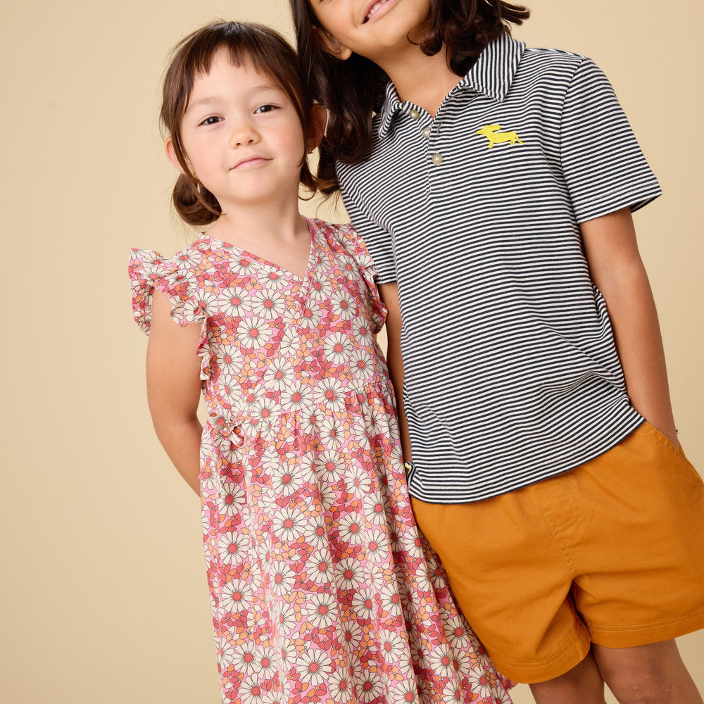 Two children, a girl in a floral dress and a boy in a striped shirt and shorts, standing against a beige background.