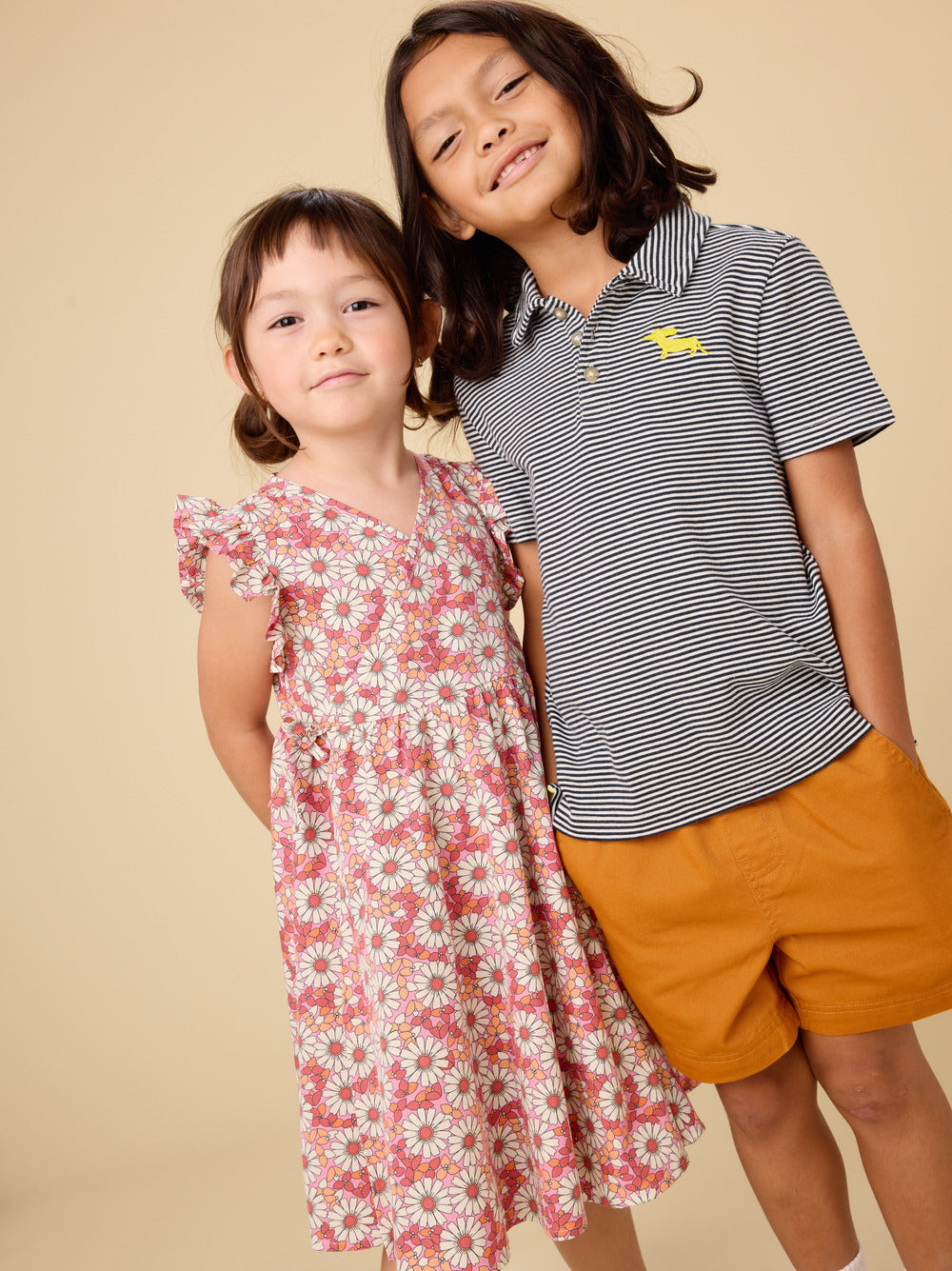 Two children, a girl in a floral dress and a boy in a striped shirt and shorts, standing against a beige background.