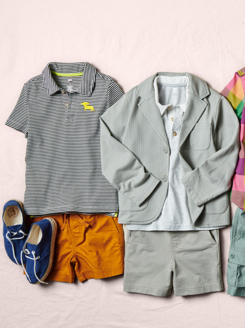 Children's clothing including a striped polo shirt, jacket, shorts, and shoes on a light pink background.