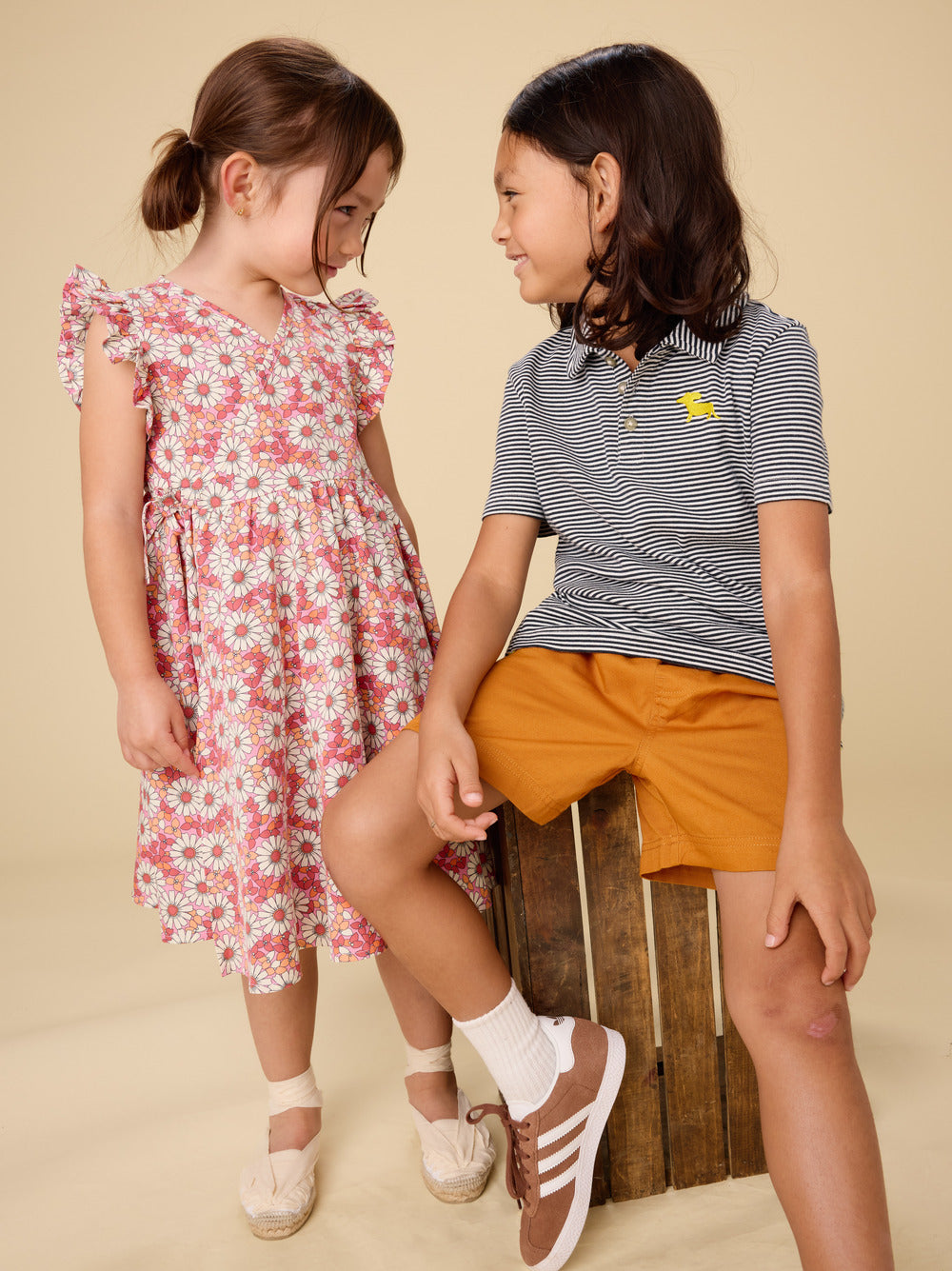 Two young girls in summer clothing sitting on a wooden crate against a beige background