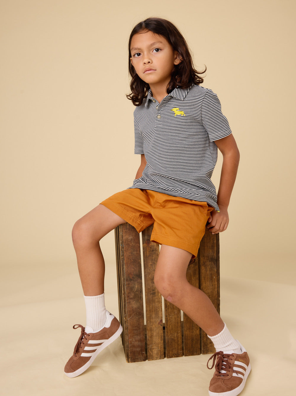 Child wearing a striped polo shirt with a logo, orange shorts, and brown shoes on a beige background