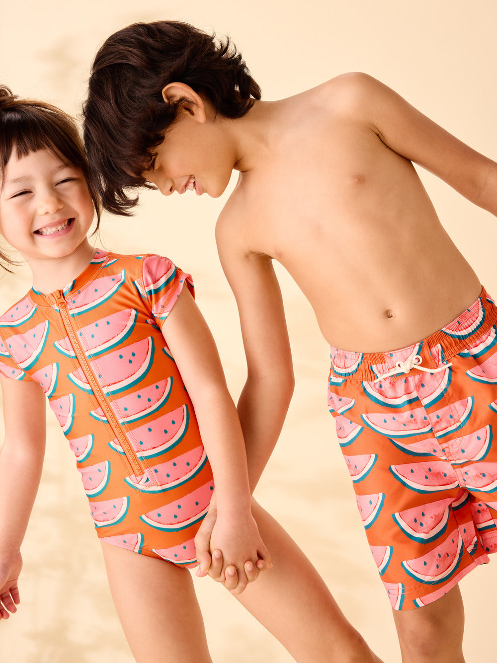 Two children wearing watermelon-patterned swimsuits on a beige background