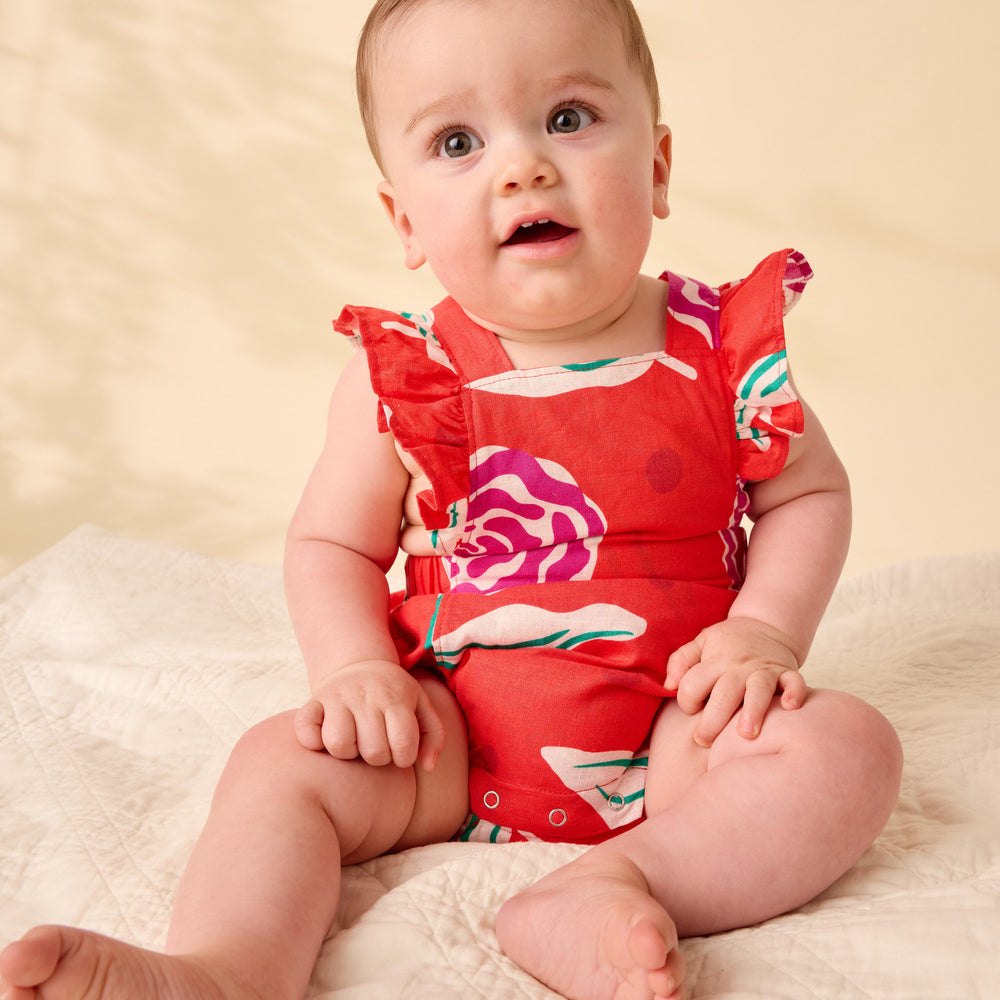 Baby wearing a red floral swimsuit sitting on a beige blanket.