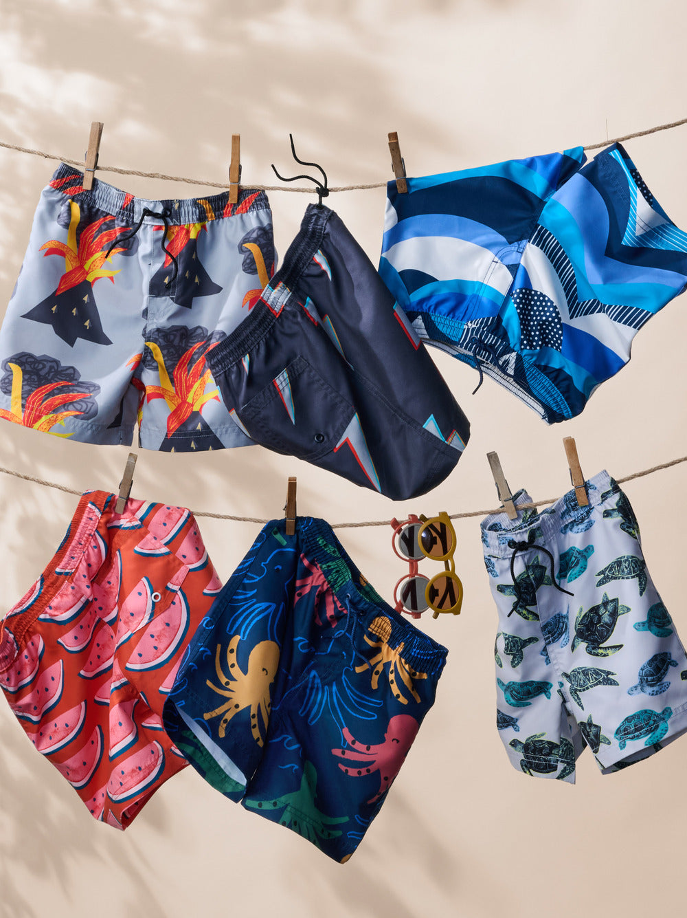 Colorful swim trunks with various patterns hanging on a clothesline against a beige background.