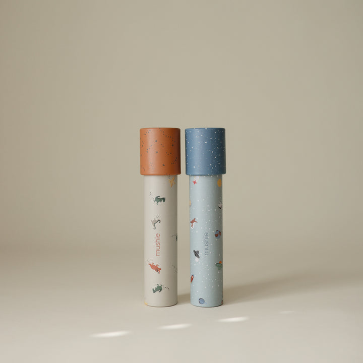 Two Mushie paper kaleidoscopes standing vertically against a light background, featuring the Space and Dinosaurs pattern with blue and orange caps.