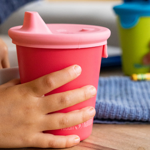 Hand holding a pink sippy cup on a wooden surface with a blurred background