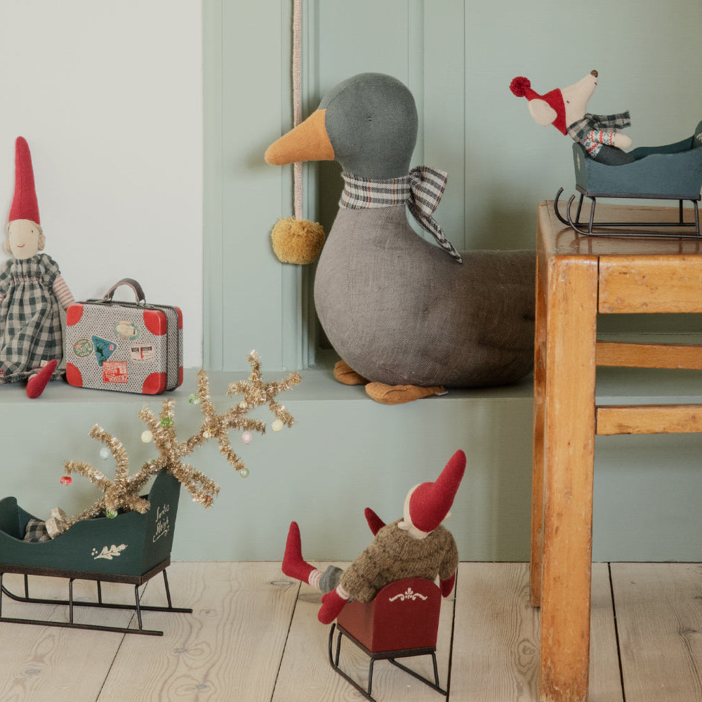 A miniature green wooden Maileg toy mouse sleigh on a wooden floor, with a larger plush toy duck, gnome dolls, a small suitcase, and holiday decor arranged in a rustic room setting.