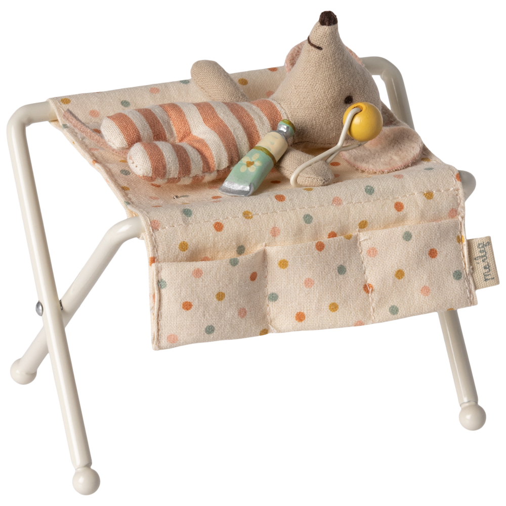 Maileg off-white miniature nursery changing table for baby mice, featuring metal folding legs, a polka-dot fabric changing pad, side pockets, a tiny rattle, and a baby lotion bottle.