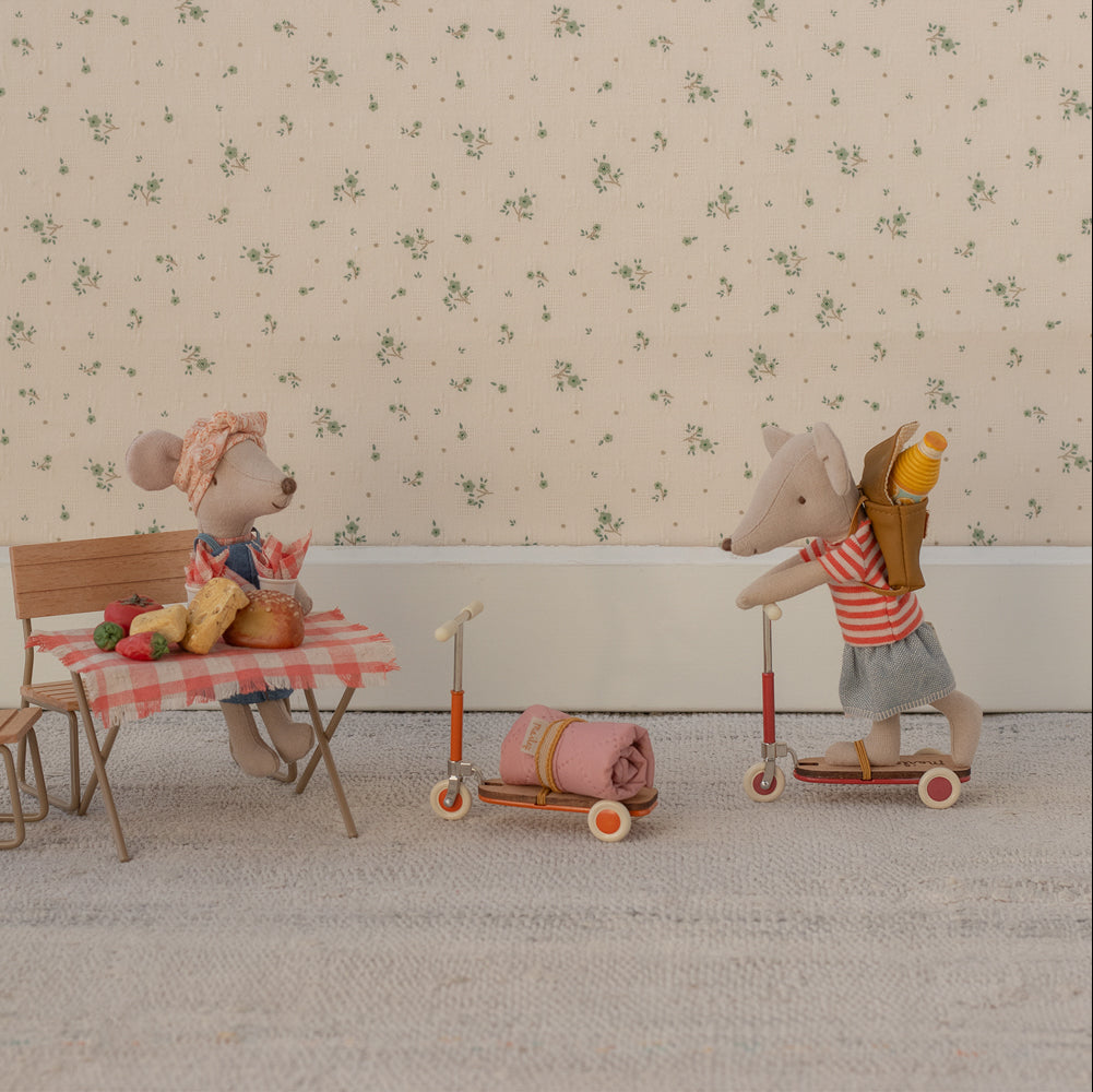 Two toy mice with a table and chairs, one on a scooter with a backpack, against a floral wallpaper background.