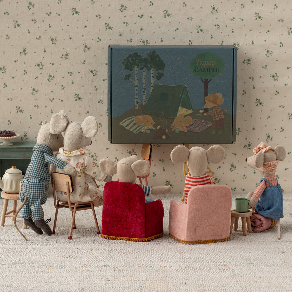 Several Maileg mouse figurines gathered in a miniature living room setup, sitting in small upholstered armchairs and chairs facing a tiny framed picture on the wall while gathered on a rug.