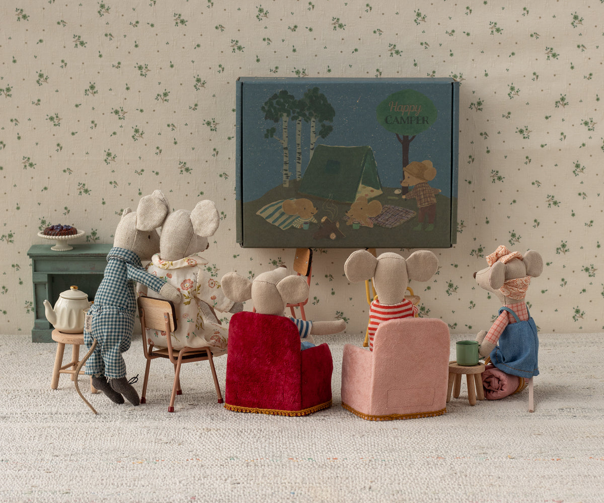 Several Maileg mouse figurines gathered in a miniature living room setup, sitting in small upholstered armchairs and chairs facing a tiny framed picture on the wall while gathered on a rug.