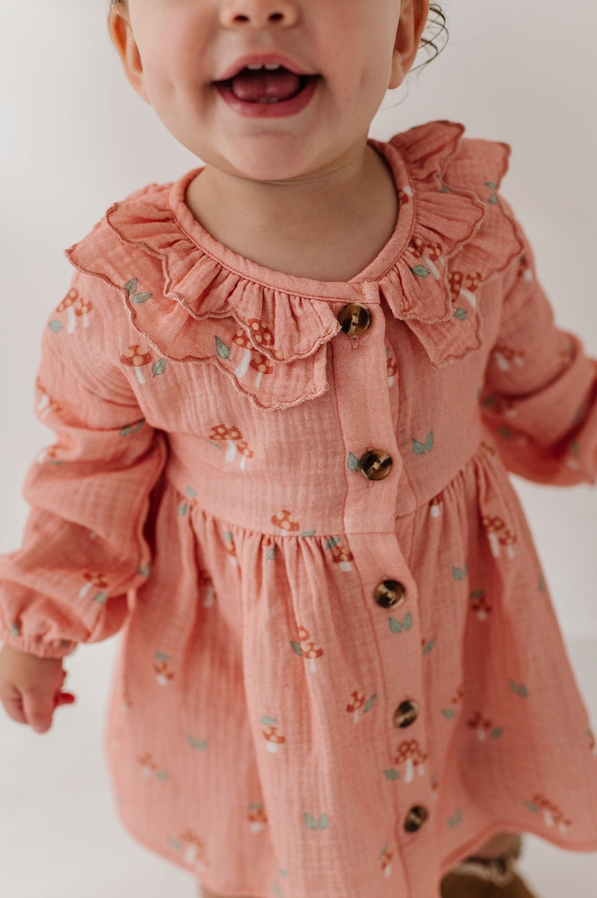 A close-up of a toddler wearing a dusty pink long-sleeve dress with a ruffled collar, button front, and a small mushroom print, against a plain light background.