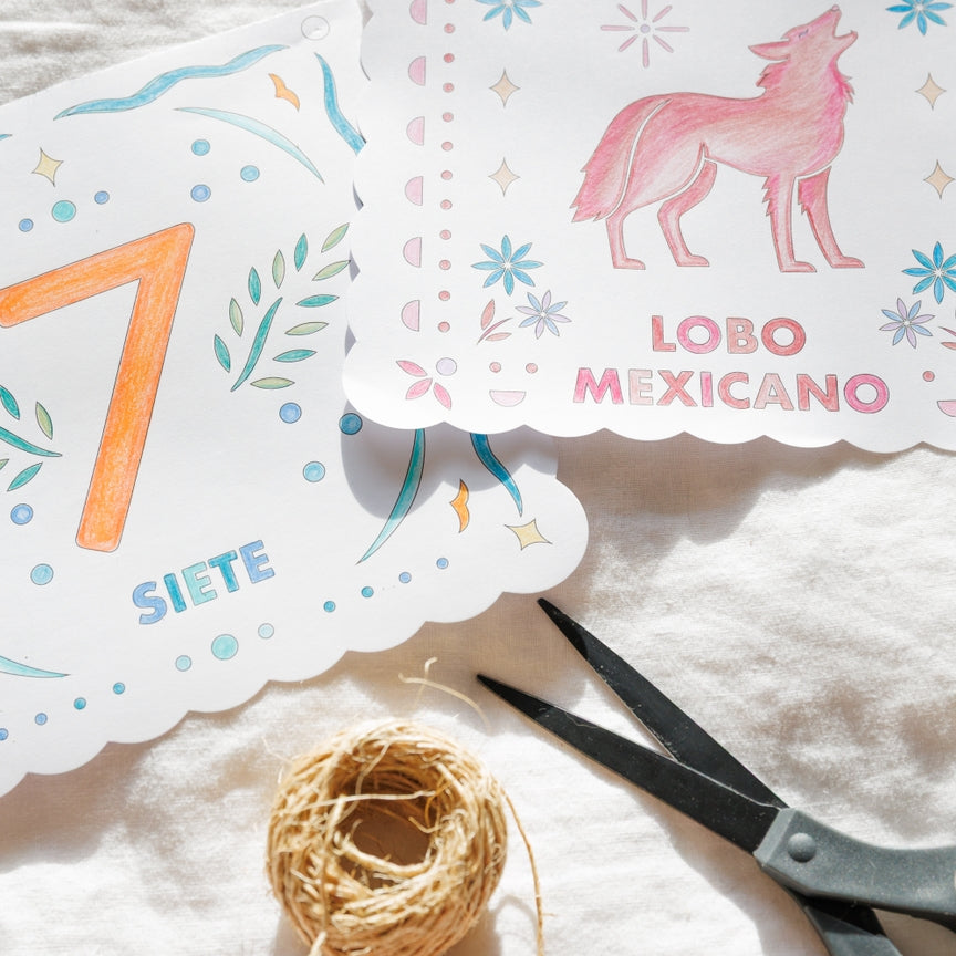 Two decorative printed paper cards with scalloped edges laid on a white fabric surface, alongside a small ball of twine and scissors. One card shows a pink wolf illustration with the text 'LOBO MEXICANO' and the letter 'L', while the other shows a large orange numeral '7' and the word 'SIETE'.