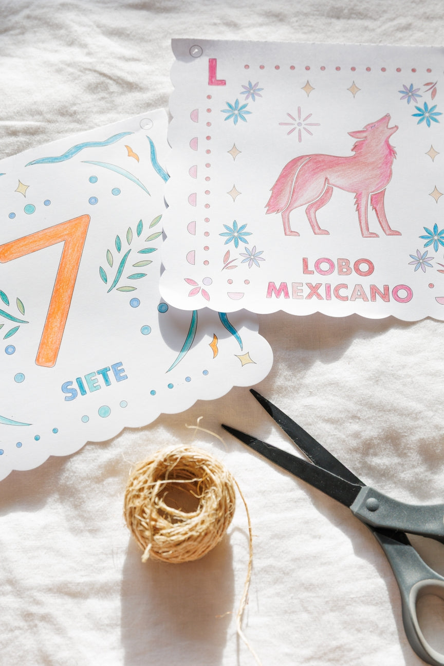 Two decorative printed paper cards with scalloped edges laid on a white fabric surface, alongside a small ball of twine and scissors. One card shows a pink wolf illustration with the text 'LOBO MEXICANO' and the letter 'L', while the other shows a large orange numeral '7' and the word 'SIETE'.