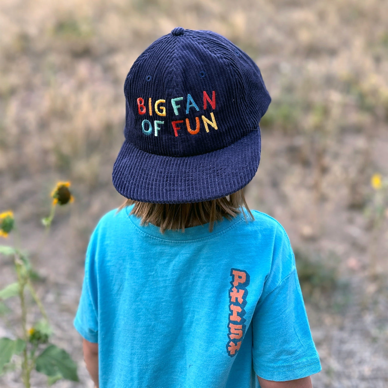 Child wearing a blue cap with colorful text and a blue shirt with red and orange text, standing in a natural setting.