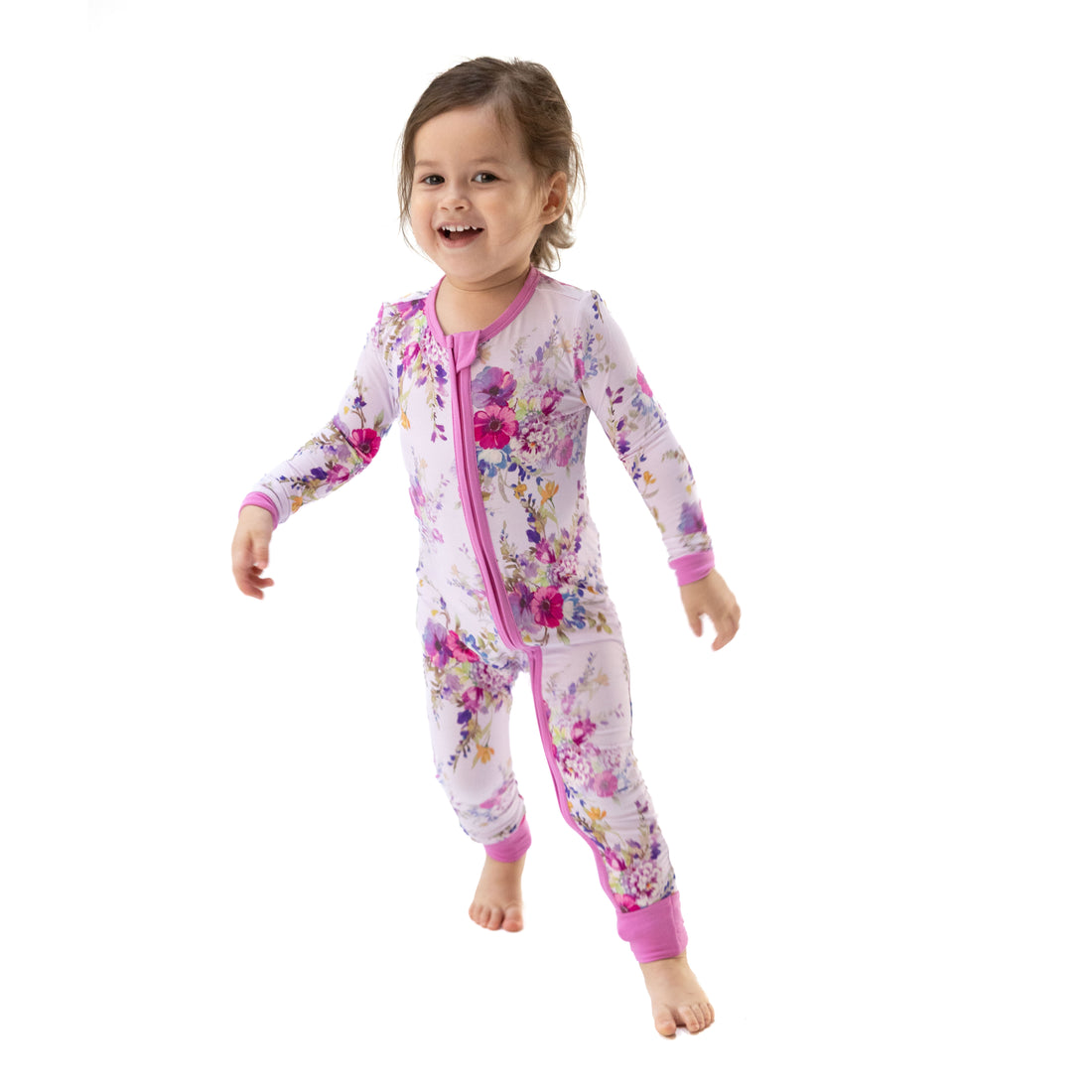A toddler girl wearing a pink and purple floral print zip-up sleeper jumpsuit, captured mid-step against a solid white background.