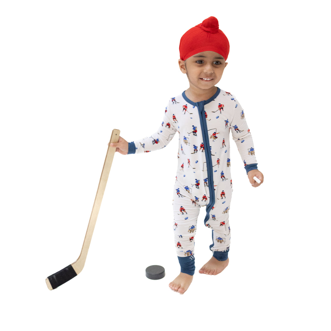 A young child wearing a red knit beanie and a white one-piece pajama with a blue zipper and blue cuffs/ankles, standing barefoot and holding a wooden toy hockey stick with a black hockey puck on a plain white background.