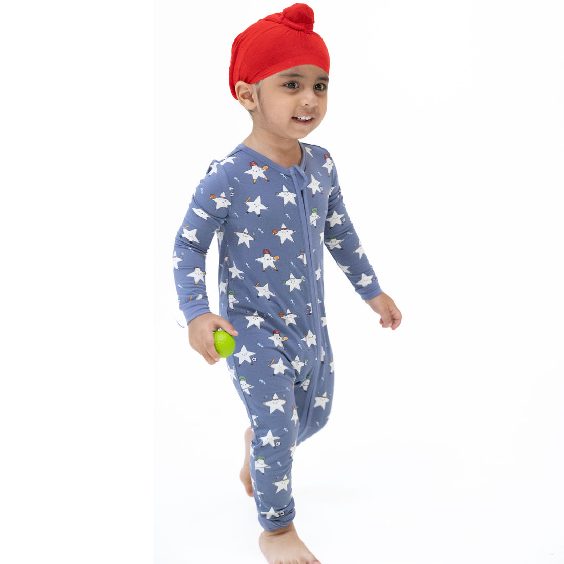 A toddler wearing a blue long-sleeve footless pajama romper with a white star pattern, a red knit beanie, and holding a small green toy, standing against a plain white background.