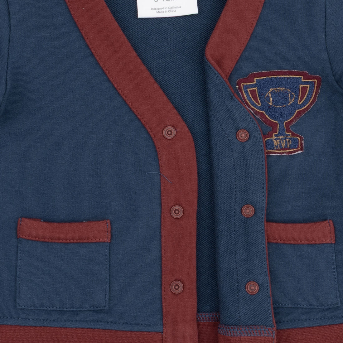 Close-up of a navy blue cardigan sweater with rust red trim, button closure, and a blue and red embroidered trophy patch on the chest.