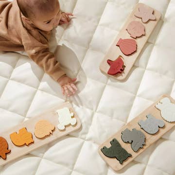 A baby in a brown outfit crawls on a white quilted mattress while playing with four wooden animal shape-sorting boards featuring different colored animal pieces.