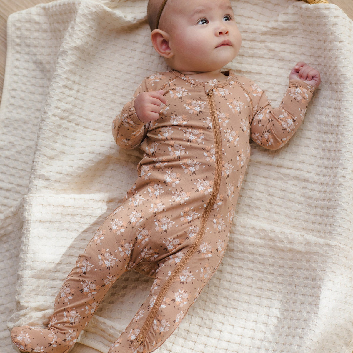 A baby lying on a cream textured blanket inside a woven bassinet, wearing a beige footed one-piece sleeper with a small white floral print and a matching headband.