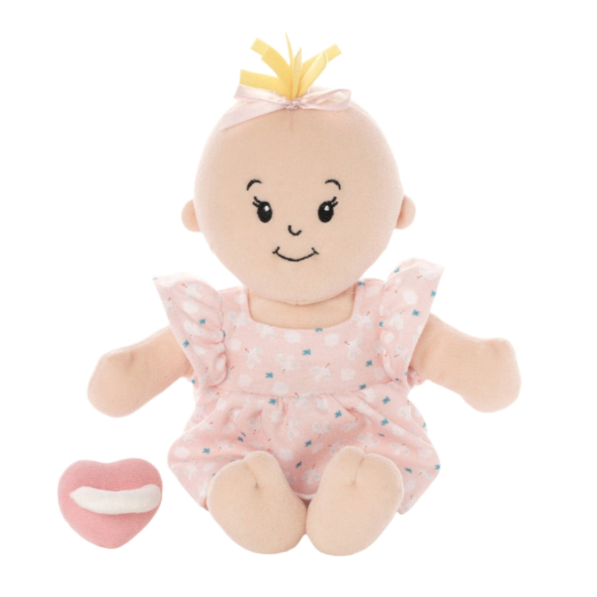A soft plush baby doll with a pale peach face and arms, wearing a light pink floral romper and a small yellow bow with pink ribbon, seated on a white background with a small pink heart toy beside it.