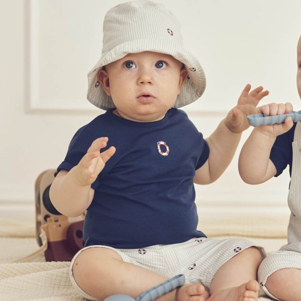 Baby wearing a navy blue shirt with a circular design and a light-colored hat, sitting on a light-colored surface.