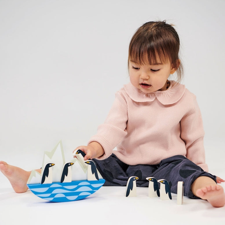 A young child with brown hair and a pink collared top sits cross-legged on the floor, carefully playing with a set of wooden balancing penguins on a rocking iceberg toy.