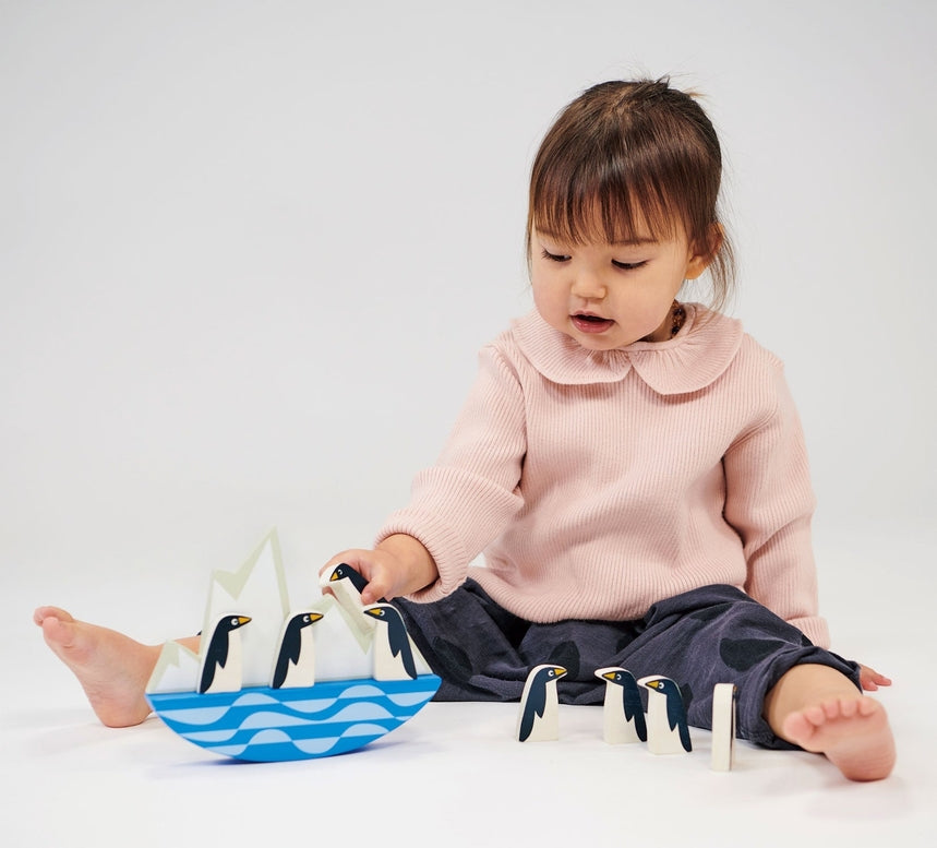 A young child with brown hair and a pink collared top sits cross-legged on the floor, carefully playing with a set of wooden balancing penguins on a rocking iceberg toy.
