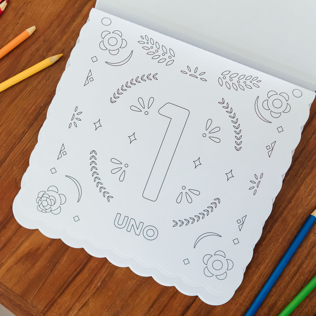 A white scalloped children's coloring book or activity sheet with the number 1 and the word UNO, photographed on a wooden table with colored pencils around it.