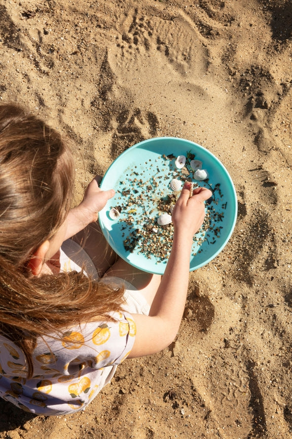 A child scooping sand into a turquoise flying disc on a beach.