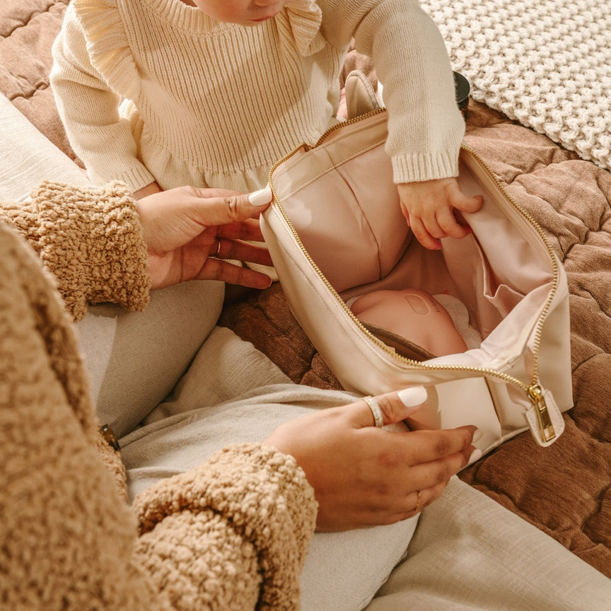 Child in a beige outfit interacting with an adult holding a pink bag on a textured surface.