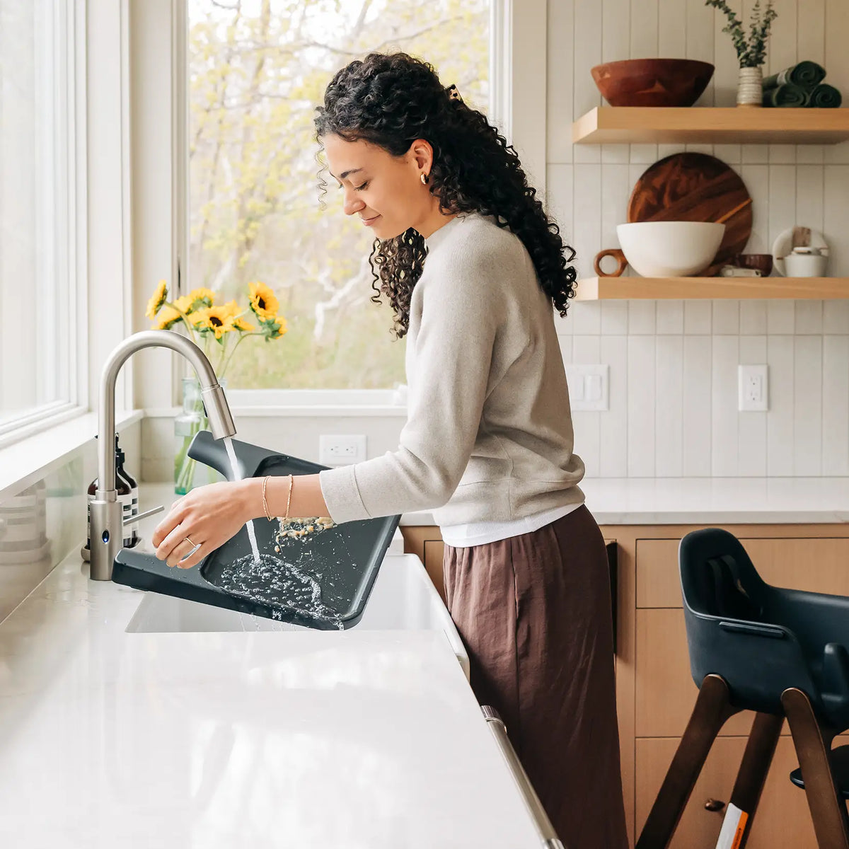 A woman with curly dark hair washes a black sink in a bright, modern kitchen with white countertops and light wood cabinets.