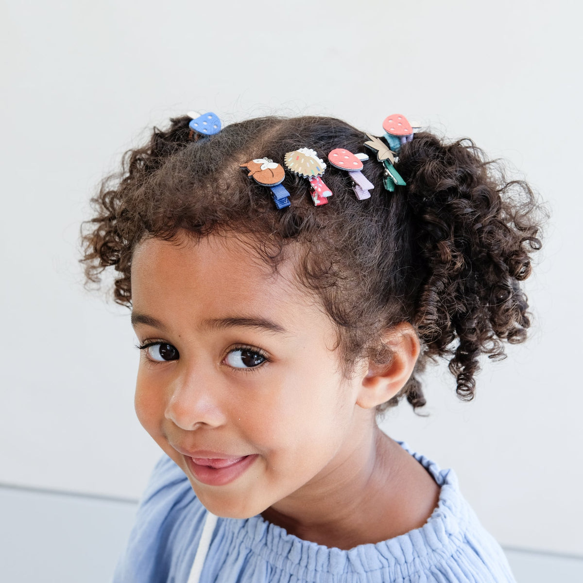 Child with curly hair adorned with colorful hair clips against a light background