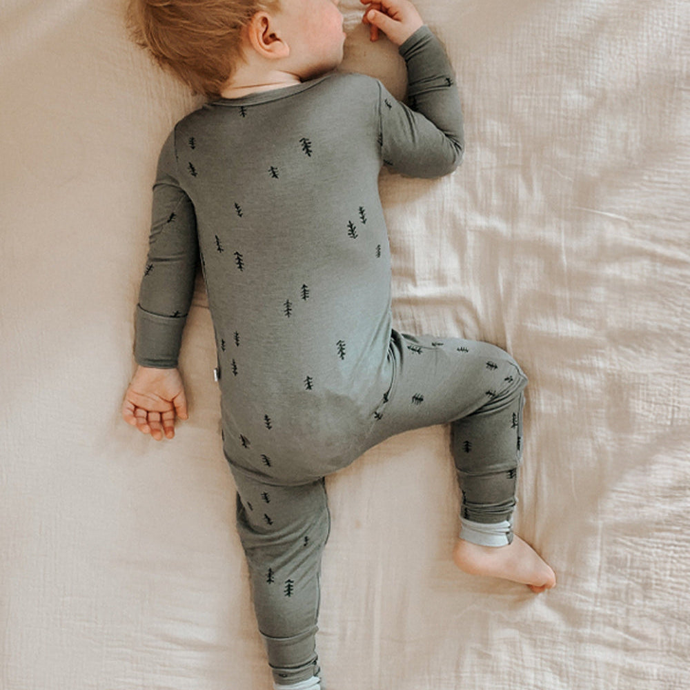 A baby lying on their side on a textured cream-colored bedsheet, wearing a sage green long-sleeve pajama romper with a small black pine tree pattern.