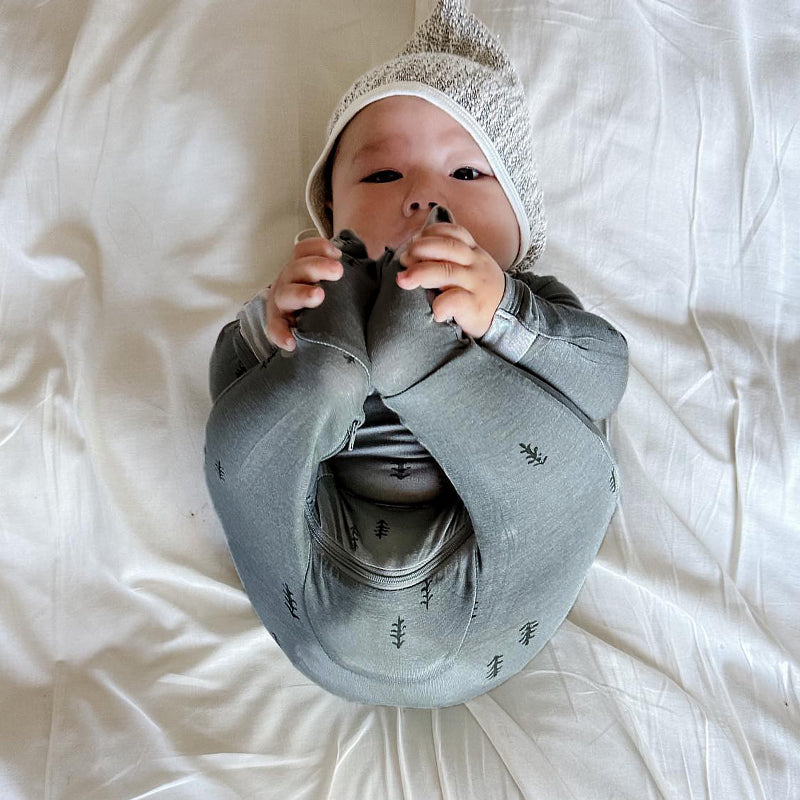 A baby lying on white bedding, wearing a gray knit huggie sleeper with small black tree prints and a matching gray knit hat.