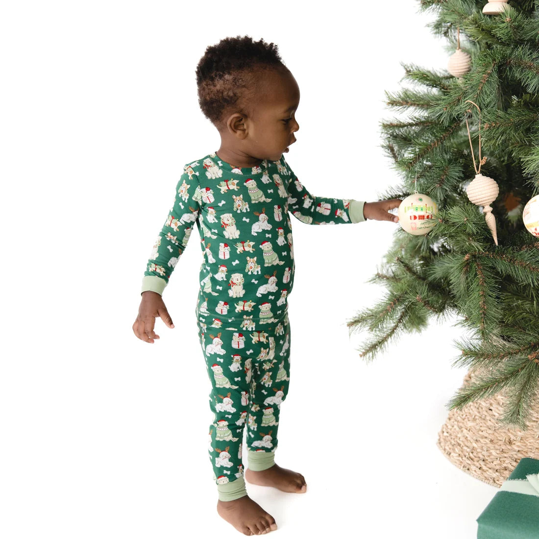 A toddler stands barefoot next to a Christmas tree, wearing a green long-sleeve pajama set with a white and light green holiday print. The background is a clean white studio setting.
