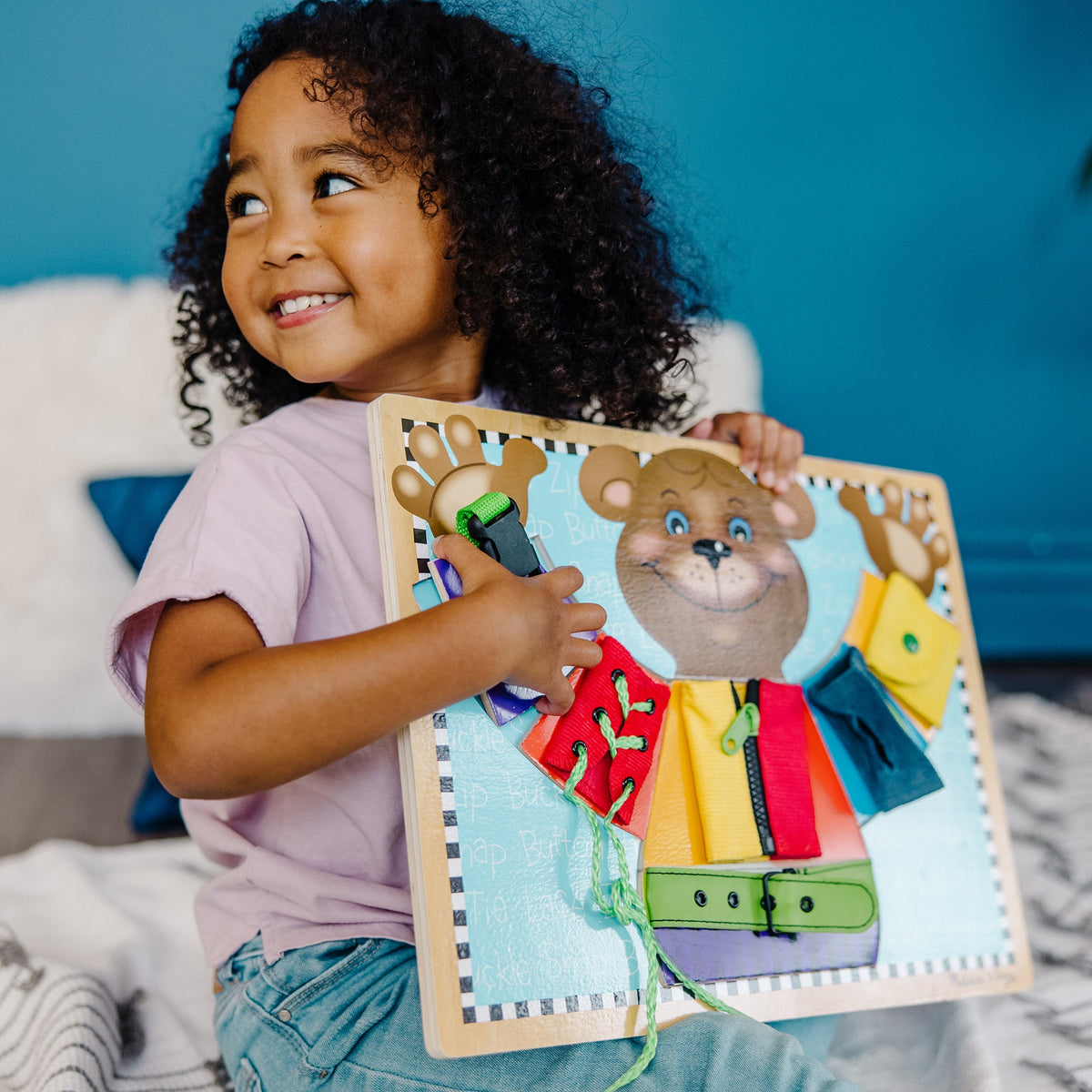 Child holding a wooden toy with a bear design against a blue wall.