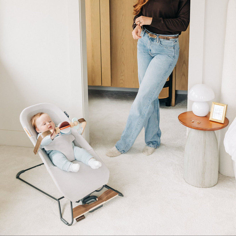 A baby sits in a light gray fabric bouncer chair with a black metal frame in a bright, modern room. A woman wearing a dark brown long-sleeve top and light blue jeans stands nearby, holding a small item.