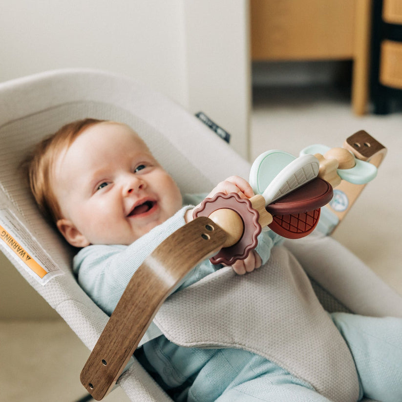 A baby wearing a light blue outfit sits in a beige baby bouncer chair, holding a wooden toy with a pink rattle.
