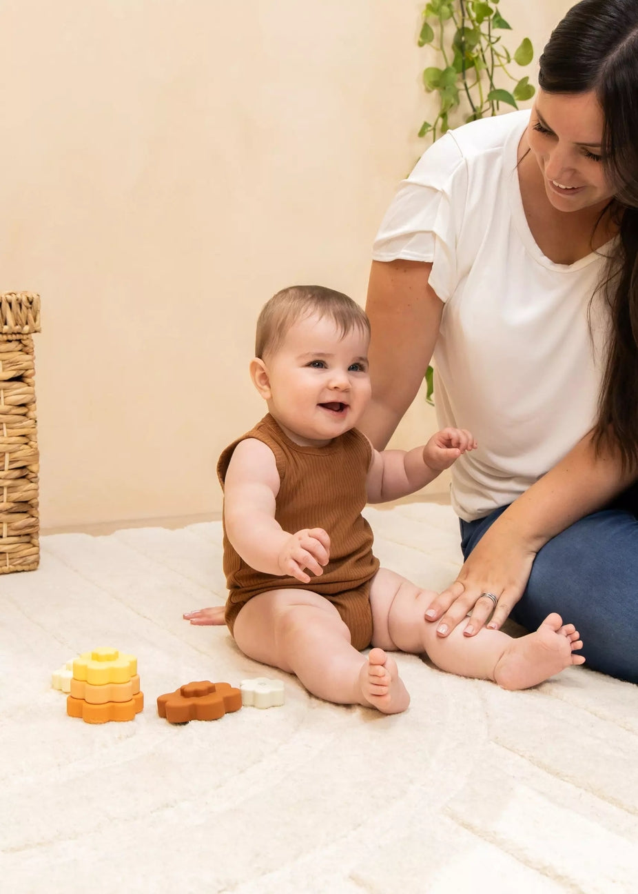 A baby wearing a brown sleeveless romper sits on a cream blanket while a woman in a white t-shirt and blue jeans kneads the baby's legs. Wooden stacking toys are on the blanket, with a wicker basket and a green plant in the background.