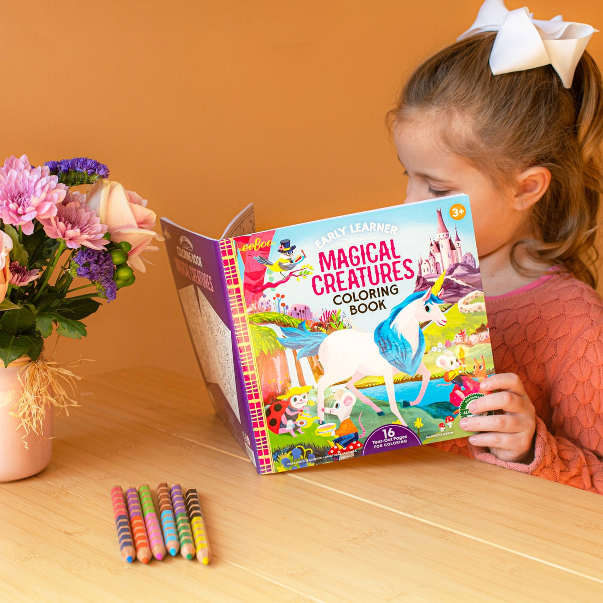 Child coloring with a 'Magical Creatures' book on a wooden table with flowers and crayons.