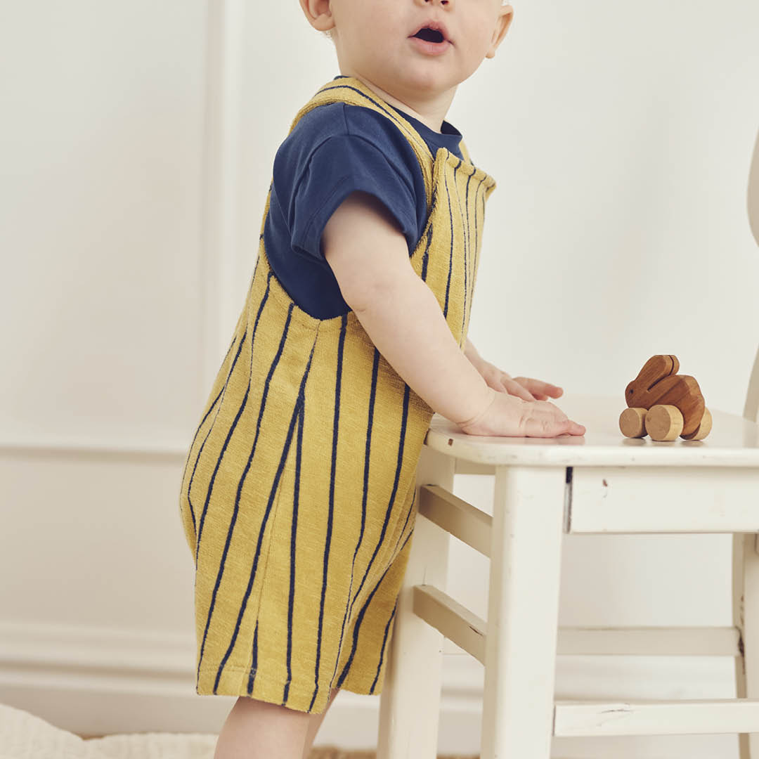 Child wearing a yellow striped dress with a blue shirt underneath, standing near a white chair.
