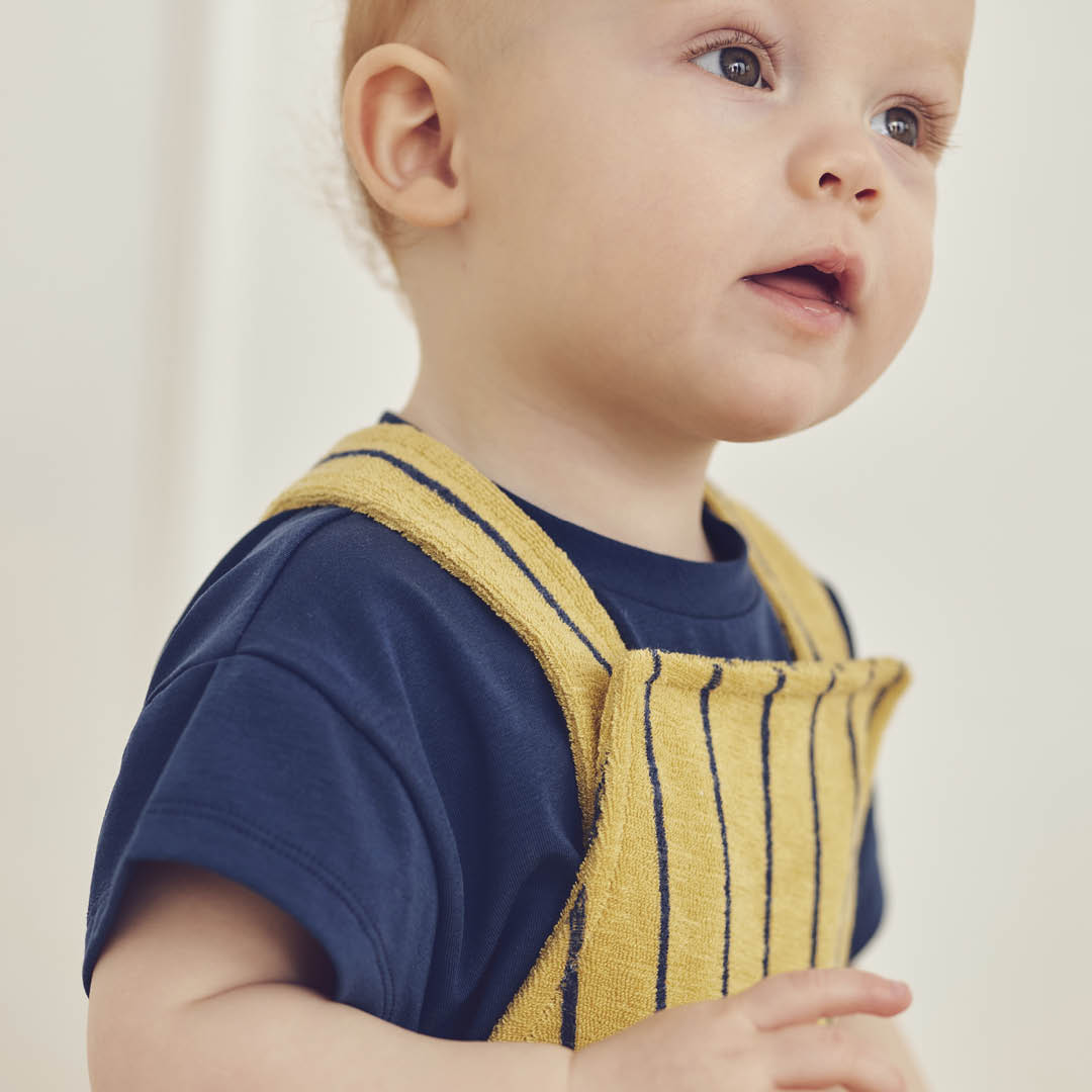 Child wearing a navy shirt and yellow striped overalls against a plain background