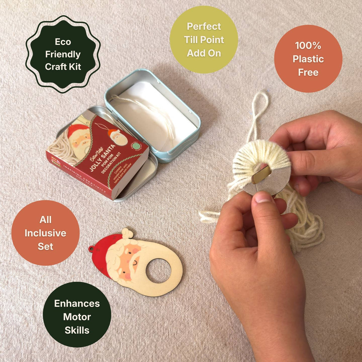 A hands-on craft activity setup on a light fabric surface, showing a person's hands wrapping white yarn around a small wooden spool. A Santa-themed wooden cutout, an open metal tin, and a Santa box are visible, along with promotional text badges including 'Eco Friendly Craft Kit,' 'Perfect Till Point Add On,' '100% Plastic Free,' 'All Inclusive Set,' and 'Enhances Motor Skills.